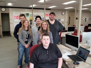 Five Lakeshore Technical College students and Dawn, the proprietor of the Red Forest Inn Bed & Breakfast, smile for a photo in a computer lab. They're surrounded by desks with computers, and a laptop showing a website, indicative of their project work for the Inn.